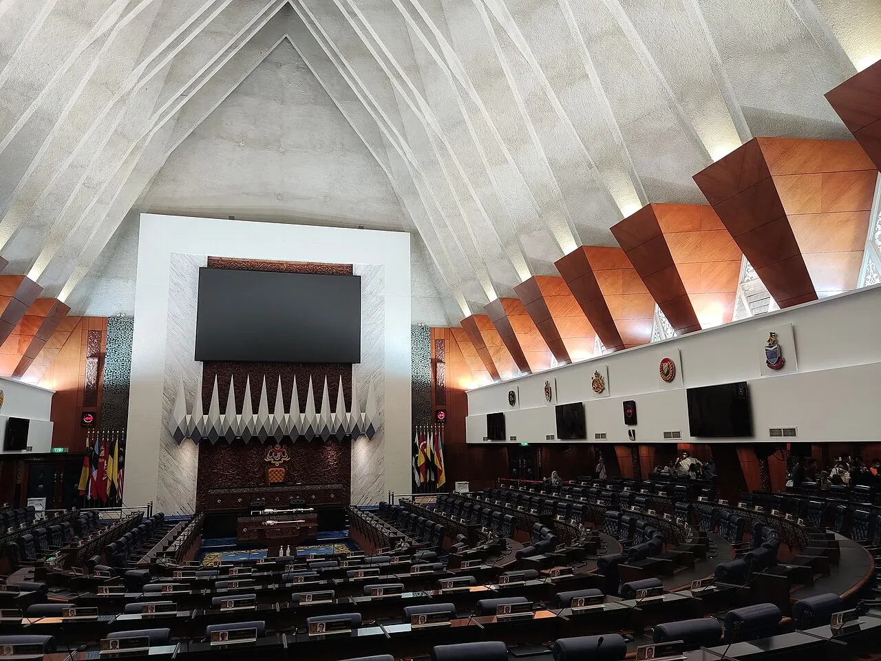 The Dewan Rakyat chamber in Malaysia's Parliament House, with rows of empty members' seats facing the Speaker's chair under a tall angled concrete roof.