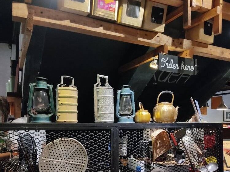 The counter of a traditional Malaysian kopitiam, with old lanterns, tiffin carriers and a brass kettle on a shelf under an 'Order here' sign
