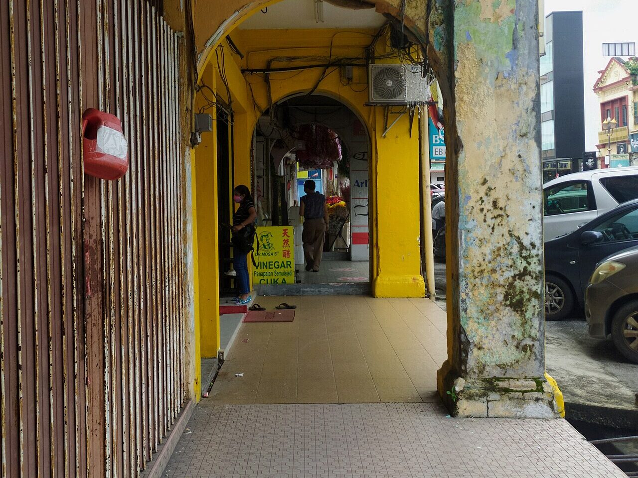 A covered five-foot walkway along a row of old shophouses in downtown Seremban, with yellow arches, a shop sign and parked cars on the street.